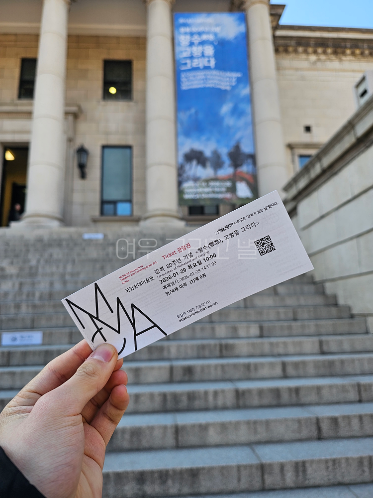 덕수궁 국립현대미술관 앞에서 건물에 걸린 전시 홍보 현수막을 배경으로 티켓을 들고 촬영한 장면